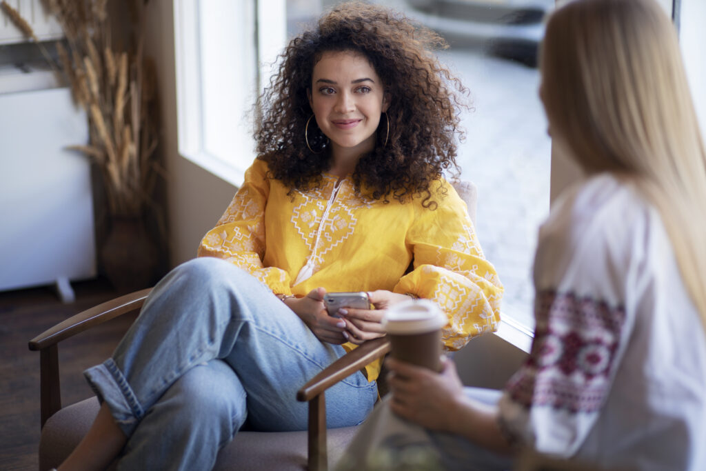 Jonge vrouw, brunette, met lang krullend haar en geel shirt aan. Ze praat met iemand die onherkenbaar in beeld is. Ze hebben allebei een koffiebeker vast.