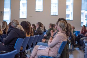 Een zaal vol mensen op stoelen naast elkaar die allemaal op links gericht zijn (je kunt niet zien waar ze naar kijken/luisteren)