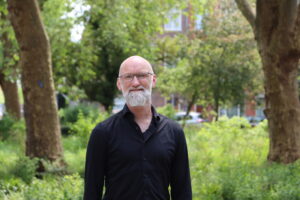 Foto van Pyschiater Jan van boven zijn middel: Een man met een kleine glimlach op zijn gezicht. Hij heeft een kaal hoofd, een rechthoekige bril op en een witte baard. Hij draagt een zwarte blouse met de bovenste knoop open. Hij staat buiten, je ziet achter hem twee boven en wat planten en nog een groot gebouw in de verte met een auto ervoor.