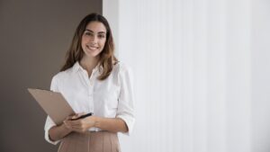 Vrouw, brunette met witte blouse en beige rok en een notitieboekje met pen in haar hand. Ze lacht. De achtergrond is ook in wit met beige. Het straalt professionaliteit uit.