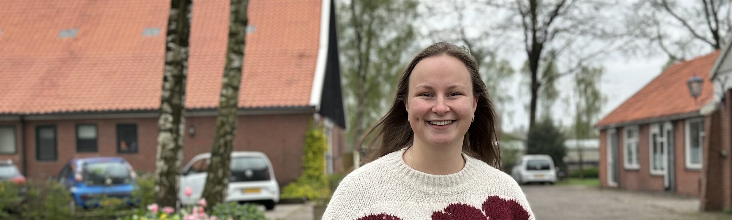 Vrouw met donkerblond haar met rode bloemetjes op witte trui. Ze staat voor de Spetse Hoeve.
