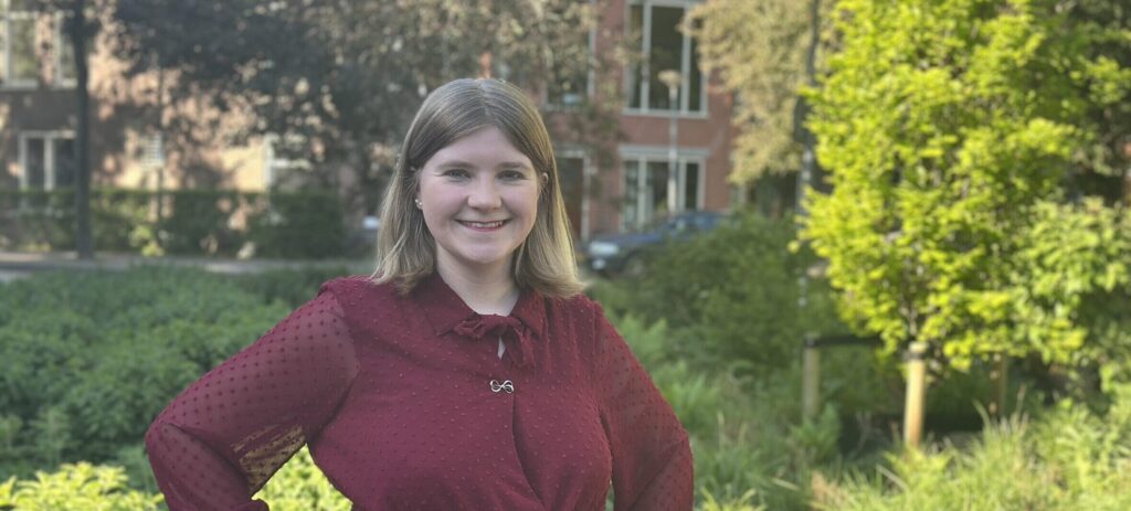 Susanne van Oosterom staat met haar handen in haar zij. Ze draagt een Donkerrode jurk en ze heeft blond haar en blauwe ogen.
