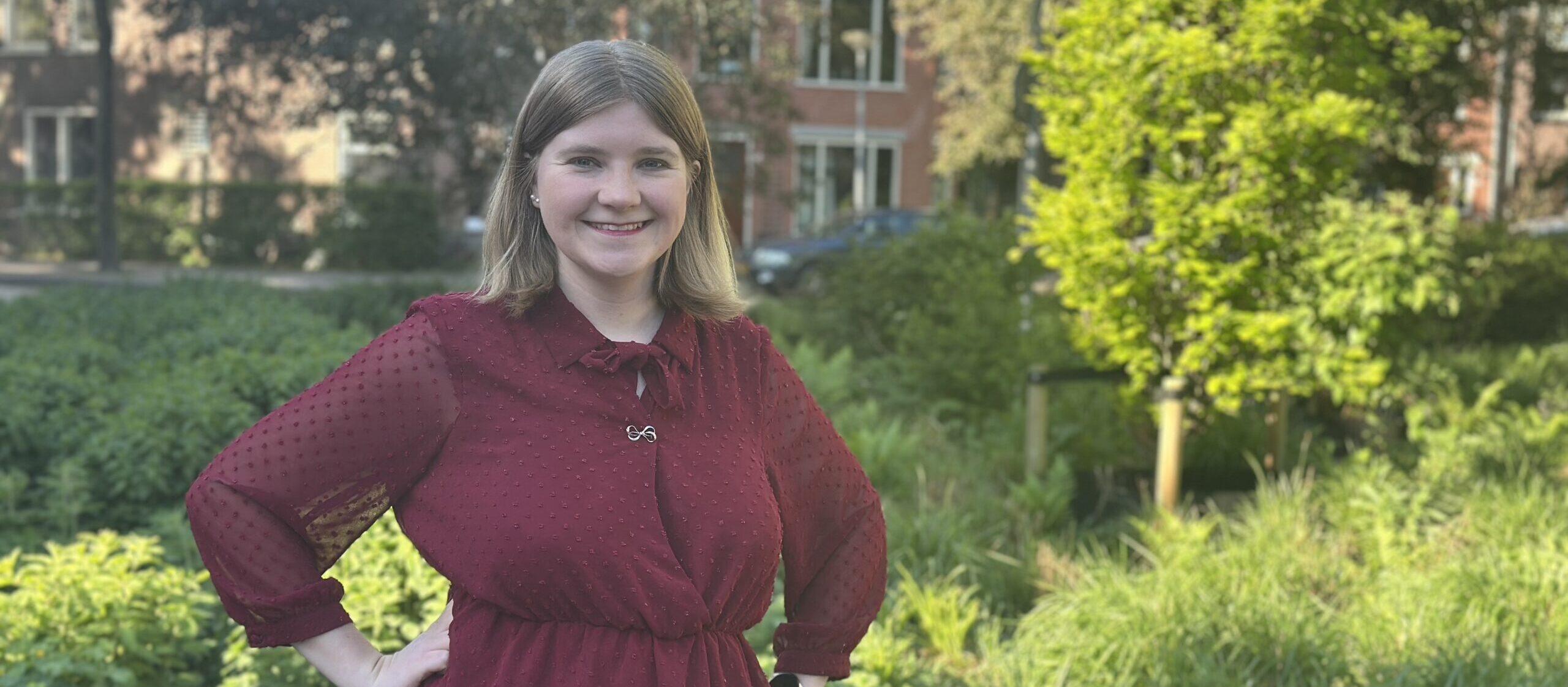 Susanne van Oosterom staat met haar handen in haar zij. Ze draagt een Donkerrode jurk en ze heeft blond haar en blauwe ogen.