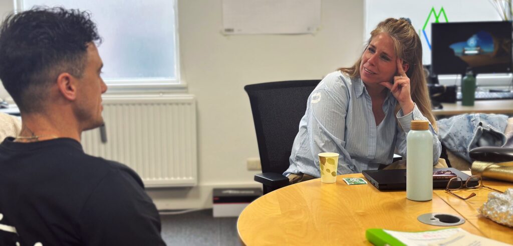 William zit links in gesprek met Janke rechts. William is een man met zwart haar en Janke heeft lang blond haar.