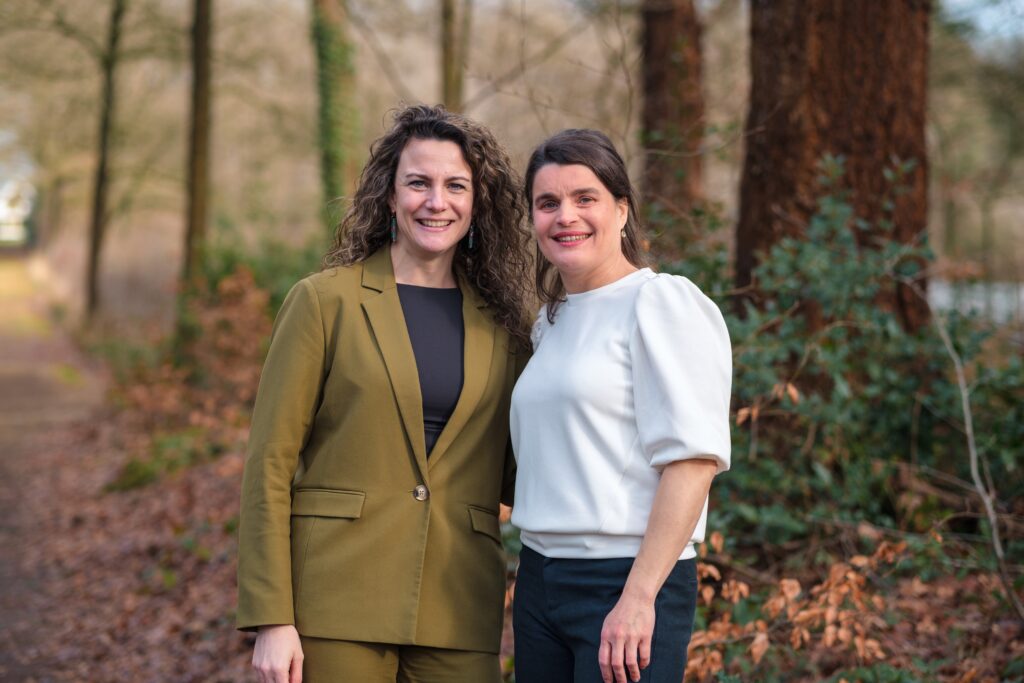 Twee vrouwelijke collega's naast elkaar met natuur op de achtergrond.